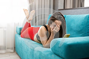 Fit brunette woman in red sport leggings and black top lying on the sofa looking upset after reading unpleasant news in her