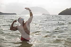 A fit and confident asian man does a classic aesthetic bodybuilding pose waist deep in the water