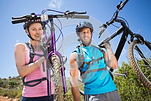 Fit couple walking down trail smiling at camera holding mountain bikes