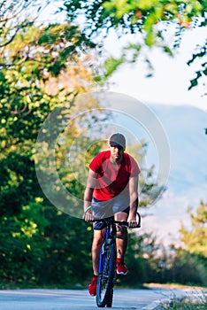 Fit cyclist rides his bicycle bike on an empty road in nature wearing a baseball hat and red t-shirt
