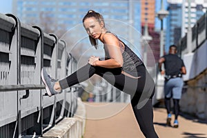 Fit female athlete runner with toned legs and butt stretches during urban city street workout, buildings in background