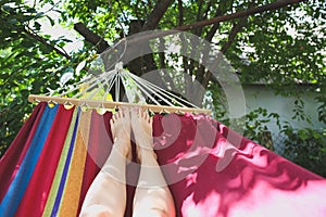 Fit girl is lying in the colourful hammock. Woman with beautiful legs is relaxing in hippie style garden near the village house.