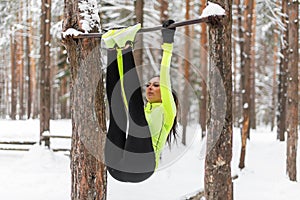 Fit girl training abs by raising legs on a horisontal bar. Fitness woman workout doing exercises outdoor winter park.