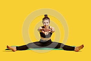 Fit gymnast woman with hair bun in tight sportswear sitting on mat with spread legs and stretching hands, warming up