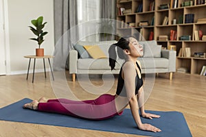 Fit korean lady doing yoga cobra pose or pilates, working out on mat in living room interior, copy space