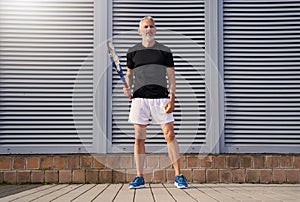 Fit mature man in sportswear holding tennis racket and looking concentrated while standing outdoors over gray background