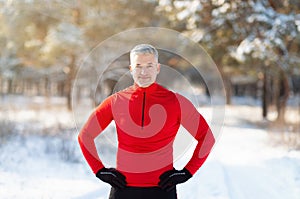 Fit mature man in sportswear standing hands on hips during his outdoor winter workout at snowy park. Seasonal sports