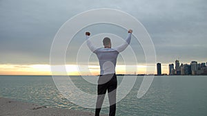 Fit muscular man goes and stops on the beach with his arms in the air. City, sea and sunrise on the background. He is a