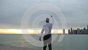 Fit muscular man goes and stops on the beach with his arms in the air. City, sea and sunrise on the background. He is a