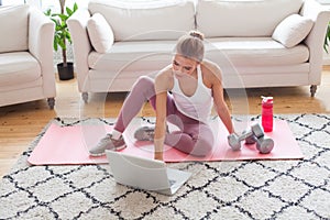 Fit pretty model in activewear sitting on mat using laptop and getting ready for a workout at home