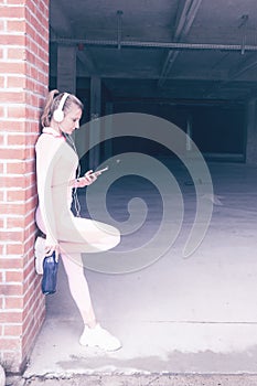 Fit pretty young woman using smartphone and headphone while resting after exercising, leaning against a brick wall