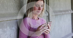 Fit pretty young woman using smartphone while resting after exercising, leaning against a brick wall outdoors