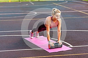 Fit sporty happy woman training on mat outdoor summer day, performing plank with foam roller massager, doing pilates