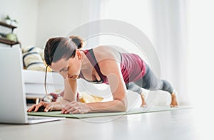 Fit sporty healthy woman on mat in low plank Chaturanga Dandasana yoga pose, doing breathing exercises, watching online yoga class