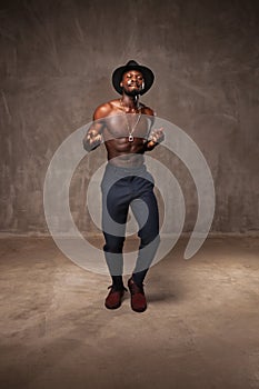 Fit strong physique African American young man wearing black hat and trousers posing dancing