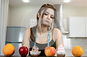 Fit woman with centimeter round neck wearing black sports top standing in the kitchen looking doubtful at cakes and fruits on the