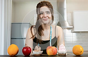 Fit woman with centimeter round neck wearing black sports top standing in the kitchen lookingat cakes and fruits on the table, photo
