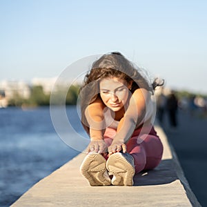 Fit woman in pink legging sitting on embankment, stretching muscles in seated forward bend position, selective focus. Hamstring