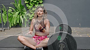 Fit woman relaxes after barbell workout in open gym, meditative pose. Athletic, toned physique showcased with healthy