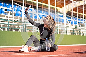 Fit woman resting after workout or running, sitting on stadium track, takes a selfie, listens to music with wired headphones on