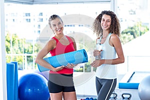 Fit women holding exercise mat and water bottle in fitness studio