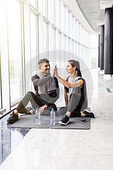 Fit young couple in sportswear smiling and high fiving together while sitting on a gym floor after working out