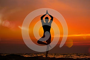 Fit young man practices vrikshasana yoga on the summer beach at sunset.