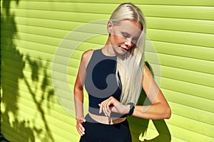 Fit young woman checking progress after workout exercises on smart watch standing against green wall outdoors. Candid portrait of