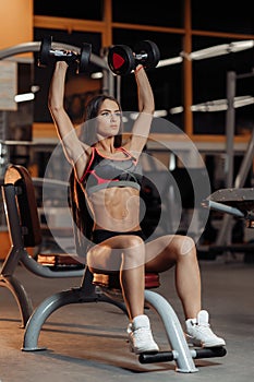 Fit young woman exercises with dumbbells for shoulders on the bench in gym.