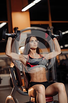 Fit young woman exercises with dumbbells for shoulders on the bench in gym.