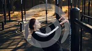 Fit young woman is exercising outdoors doing pull-ups on low horizontal bar in park in autumn morning training alone