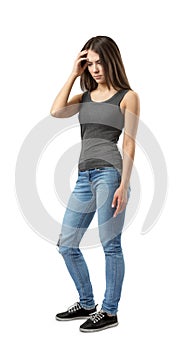 Fit young woman in gray sleeveless top and blue jeans standing in half-turn with troubled look on face and one hand on