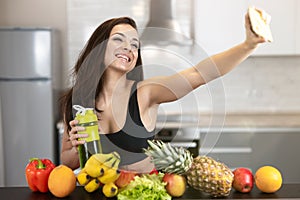 Fit young woman wearing sexy black sports top shooting selfie standing in the kitchen full of fruits, dietology and nutrition