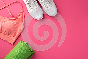 Fitness accessories concept. Top view photo of green yoga mat pink sports top and white shoes on isolated pink background with