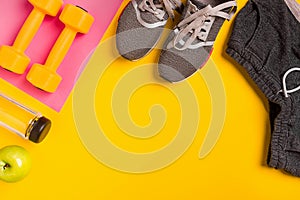 Fitness accessories on yellow background. Sneakers, bottle of water and dumbbells