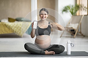 Fitness App. Pregnant Woman Sitting On Yoga Mat And Showing Blank Smartphone