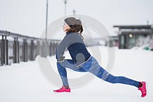 Fitness athlete female model runner doing flexibility exercise for legs before run at snow winter promenade