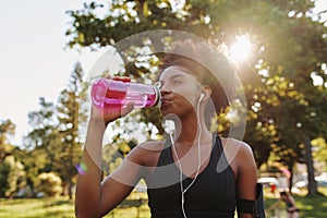 Fitness athlete young african american woman listening to music on earphones drinking water in a reusable water bottle