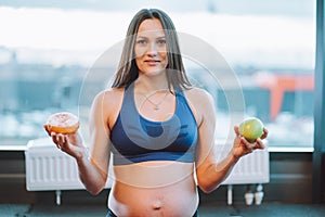 Fitness attractive pregnant woman in sportswear. Doing yoga exercise, choise green apple, donut. Diet starving. Close up