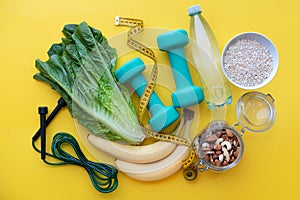 Fitness background with water, apple, dumbbells, nuts, and tape measure on yellow background.