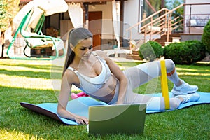 Fitness beautiful slim woman doing side plank with resistance band and watching online tutorials on laptop, training