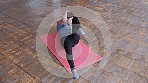 Fitness body flex. The athletic girl in a gray T-shirt and black tights lies on a pink rug and alternately bends her