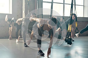 Fitness Center. Portrait of men and women doing fitness training exercises at industrial gym. Straps, group workout