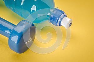 Fitness concept with dumbbell and bottle/blue dumbbell and bottle on a yellow background. Top view