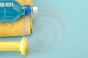 Fitness concept with dumbbell, towel and bottle/yellow dumbbell, towel and bottle on a blue background. Copy space. Top view