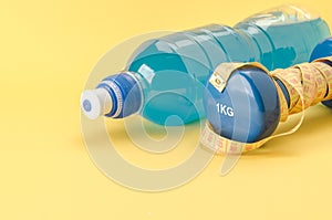 Fitness concept with dumbbell with yellow centimeter and bottle/blue dumbbell with centimeter and bottle on a yellow background.