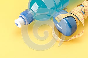 Fitness concept with dumbbell with yellow centimeter and bottle/blue dumbbell with centimeter and bottle on a yellow background.