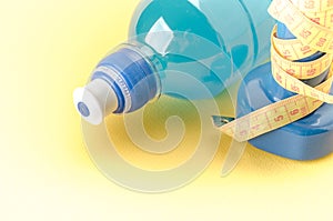 Fitness concept with dumbbell with yellow centimeter and bottle/blue dumbbell with centimeter and bottle on a yellow background.