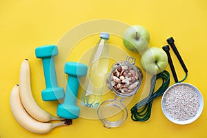 Fitness concept - fruits, water, nuts, dumbbells and skipping rope on yellow background.