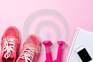 Fitness concept, pink sneakers and dumbbells with notebook with smart phone on pink background, top view with copy space